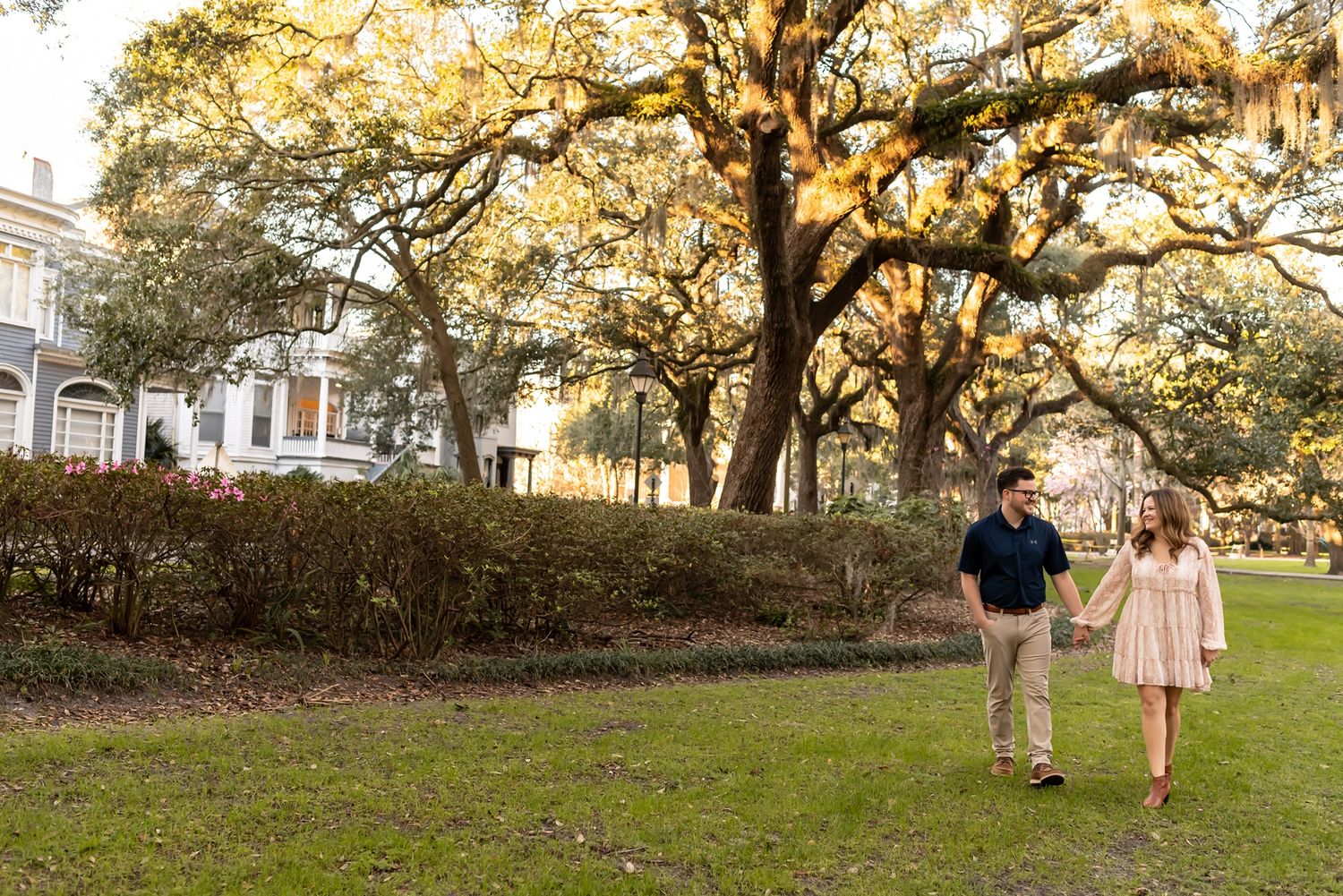 Couple walking hand in hand under Spanish moss draped oak trees in a historic southern garden setting at sunset.