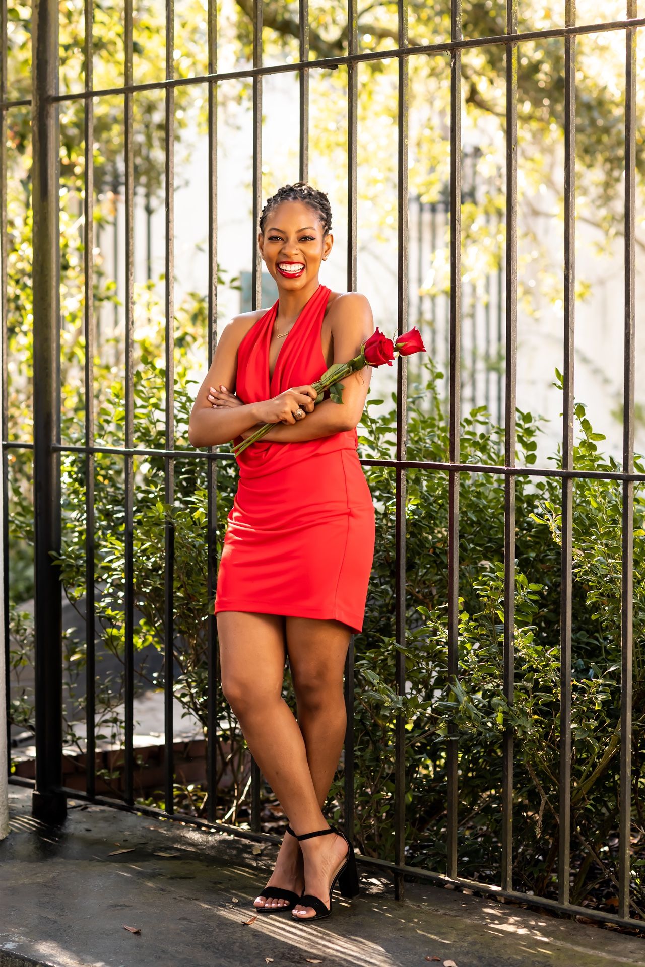 A person in a vibrant red dress poses confidently against an iron fence with greenery in the background.