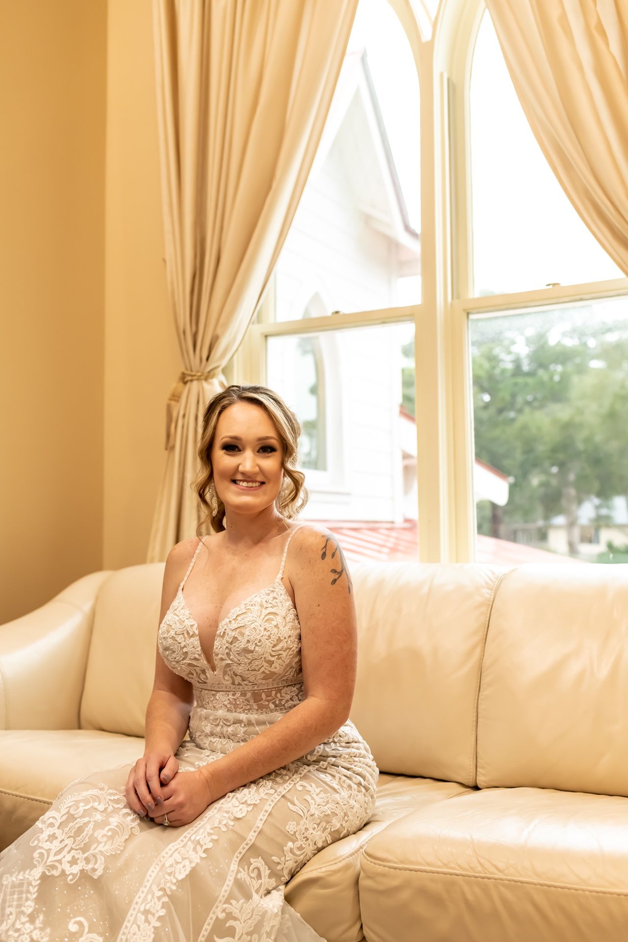 A bride in an ornate lace wedding dress sits elegantly on a cream leather couch by a tall window with beige curtains.
