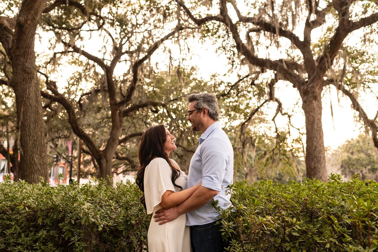 Couple embraces in golden sunlit garden surrounded by oak trees during romantic engagement photo session.