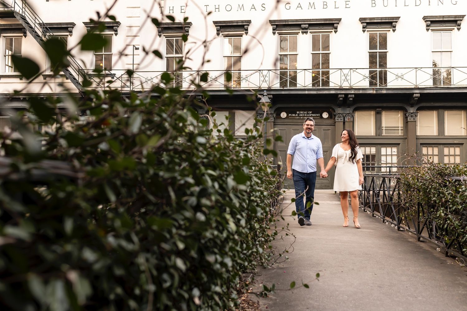 A couple walks along a garden pathway beside a historic building with tall windows and hedges.
