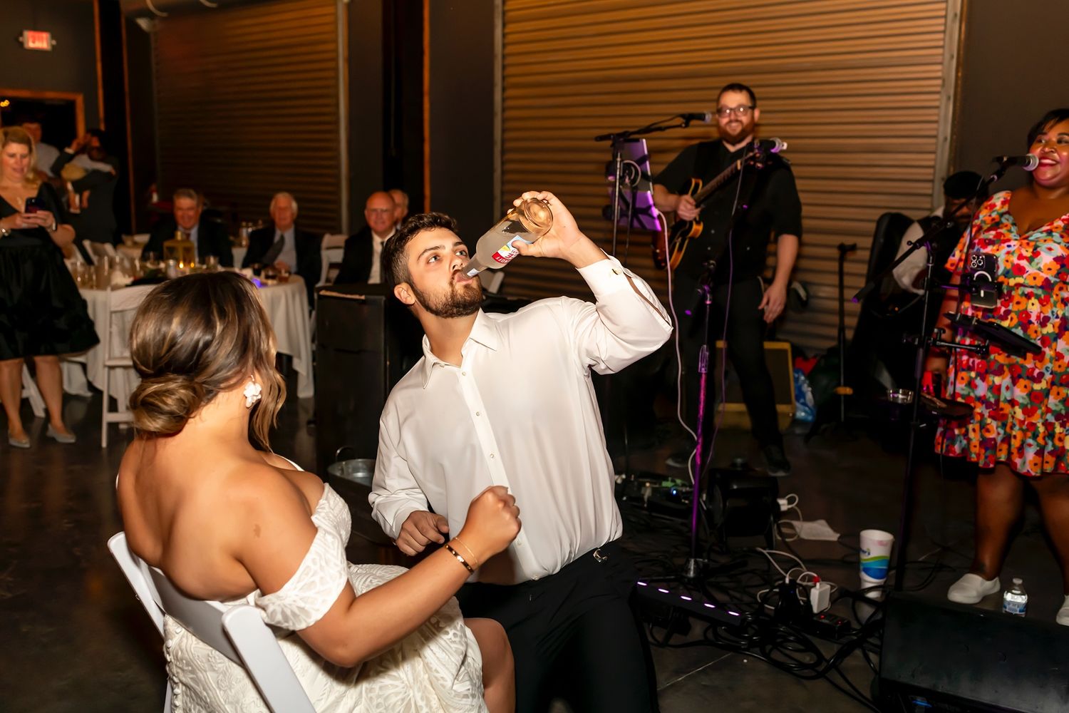 A bride and groom share a moment at their wedding reception as the groom drinks from a bottle while wedding guests watch.