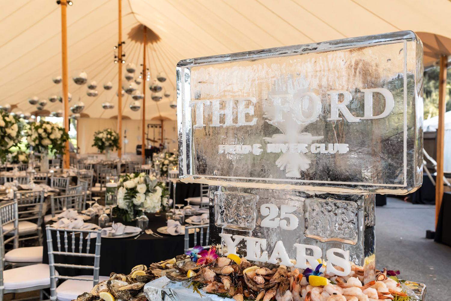 Wedding reception table setup with an elegant ice sculpture displaying The Ford logo under a white tent canopy.
