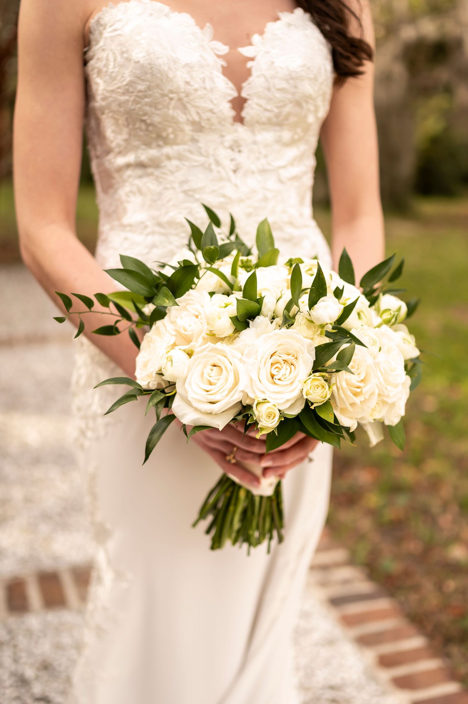 A bride in a lace wedding dress holds an elegant white rose bouquet with greenery against a garden pathway background.