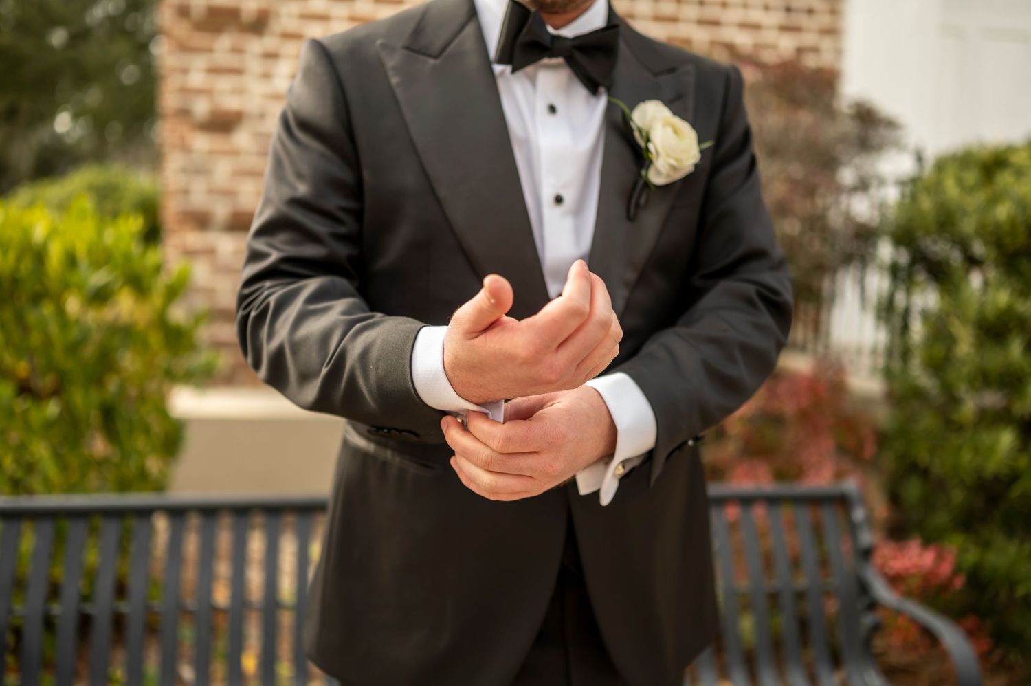 A groom in a black tuxedo and bowtie adjusts his cuffs while wearing a white boutonniere on his lapel.