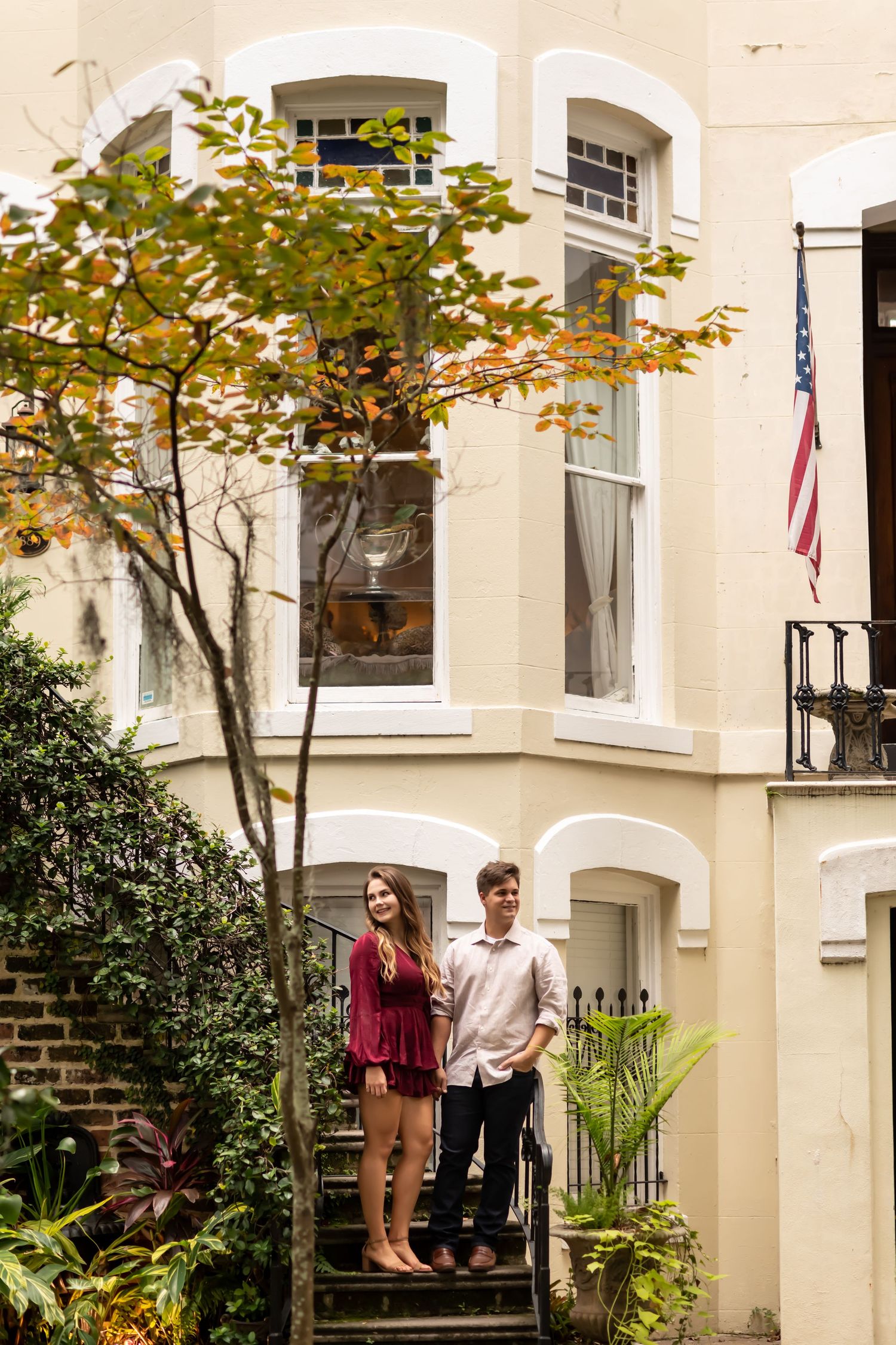 A couple stands on the steps of a classic Charleston-style home with a white facade and American flag in the garden.