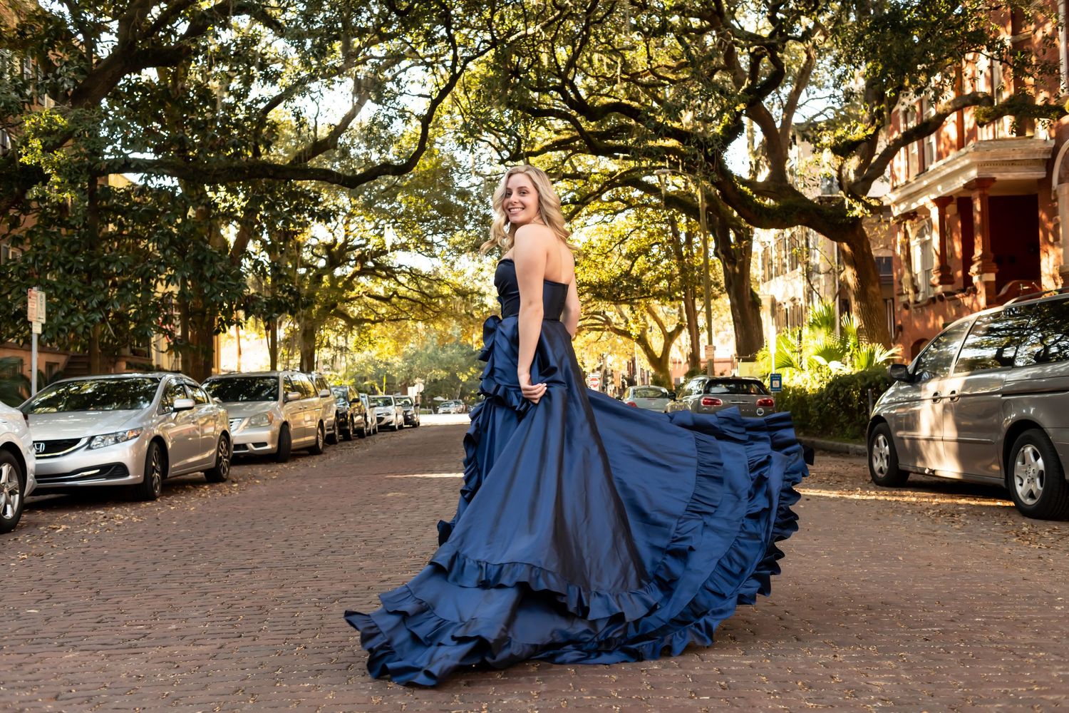 Person in a flowing navy blue ball gown twirls on a tree-lined street with parked cars and historic architecture.