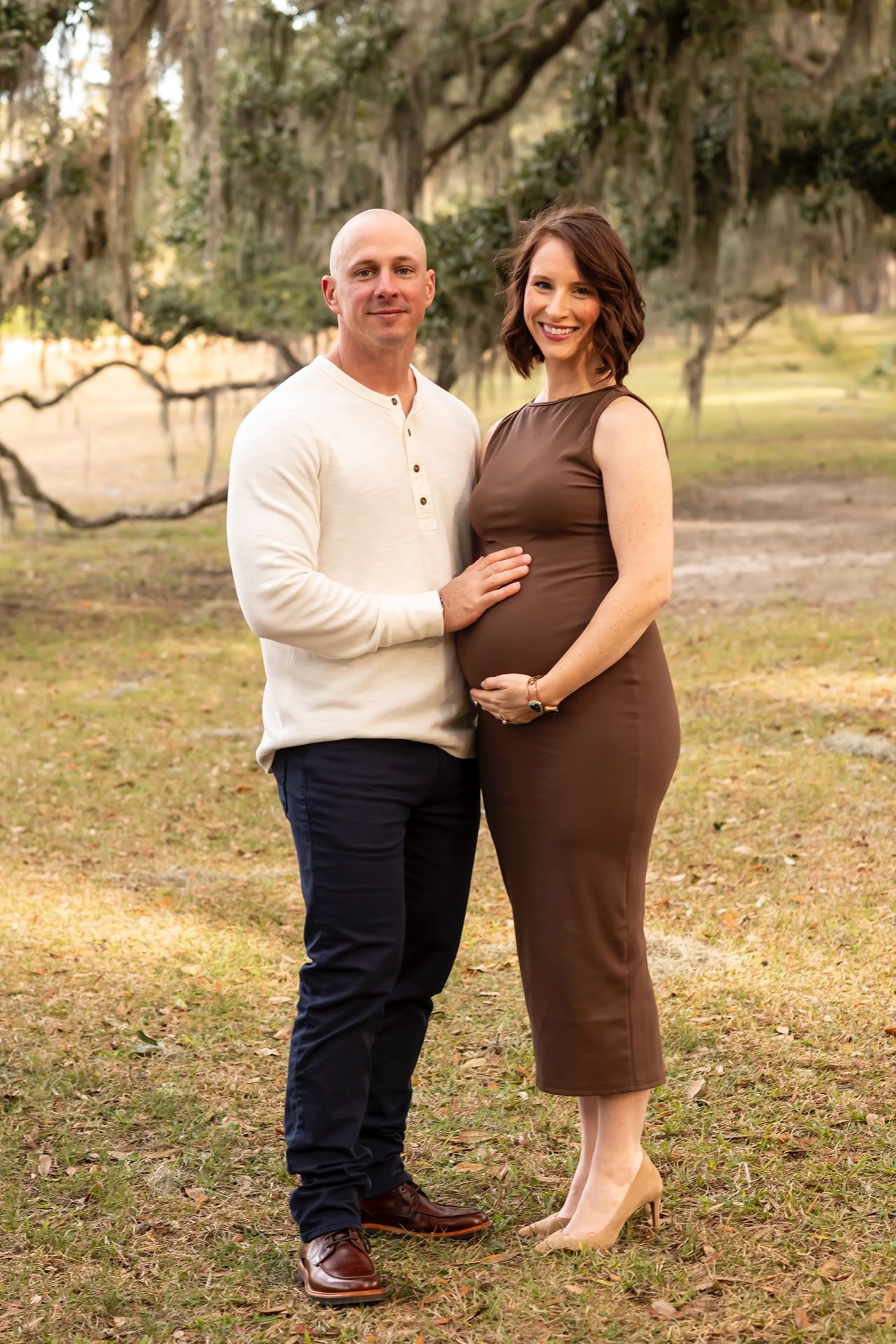 A couple poses together on grass under Spanish moss-draped trees at sunset in a romantic outdoor setting.