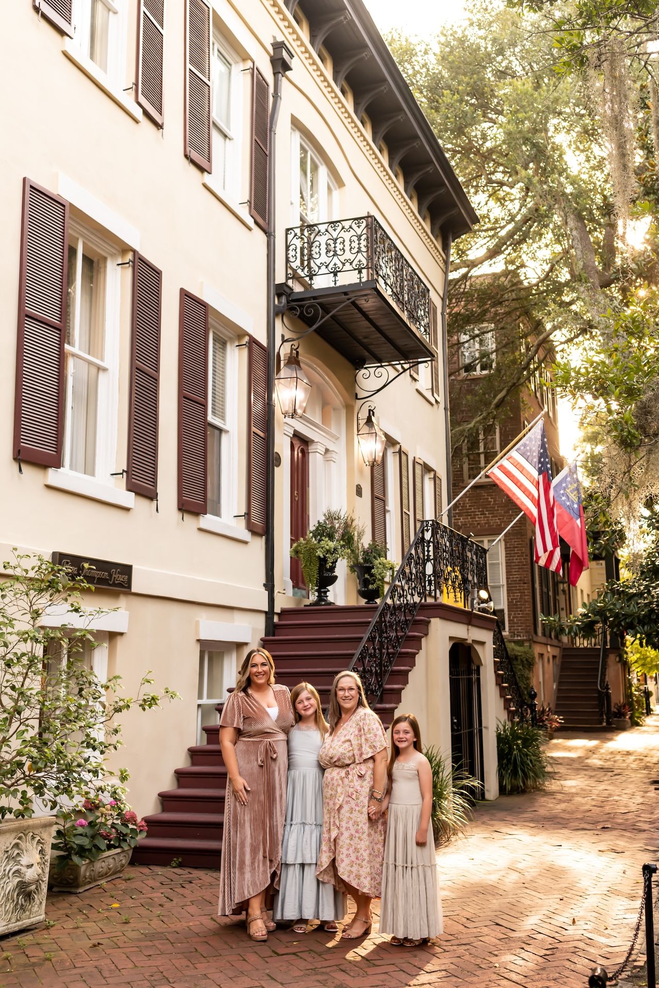 Four women in pastel dresses pose on a brick path outside a historic Southern home with an American flag.