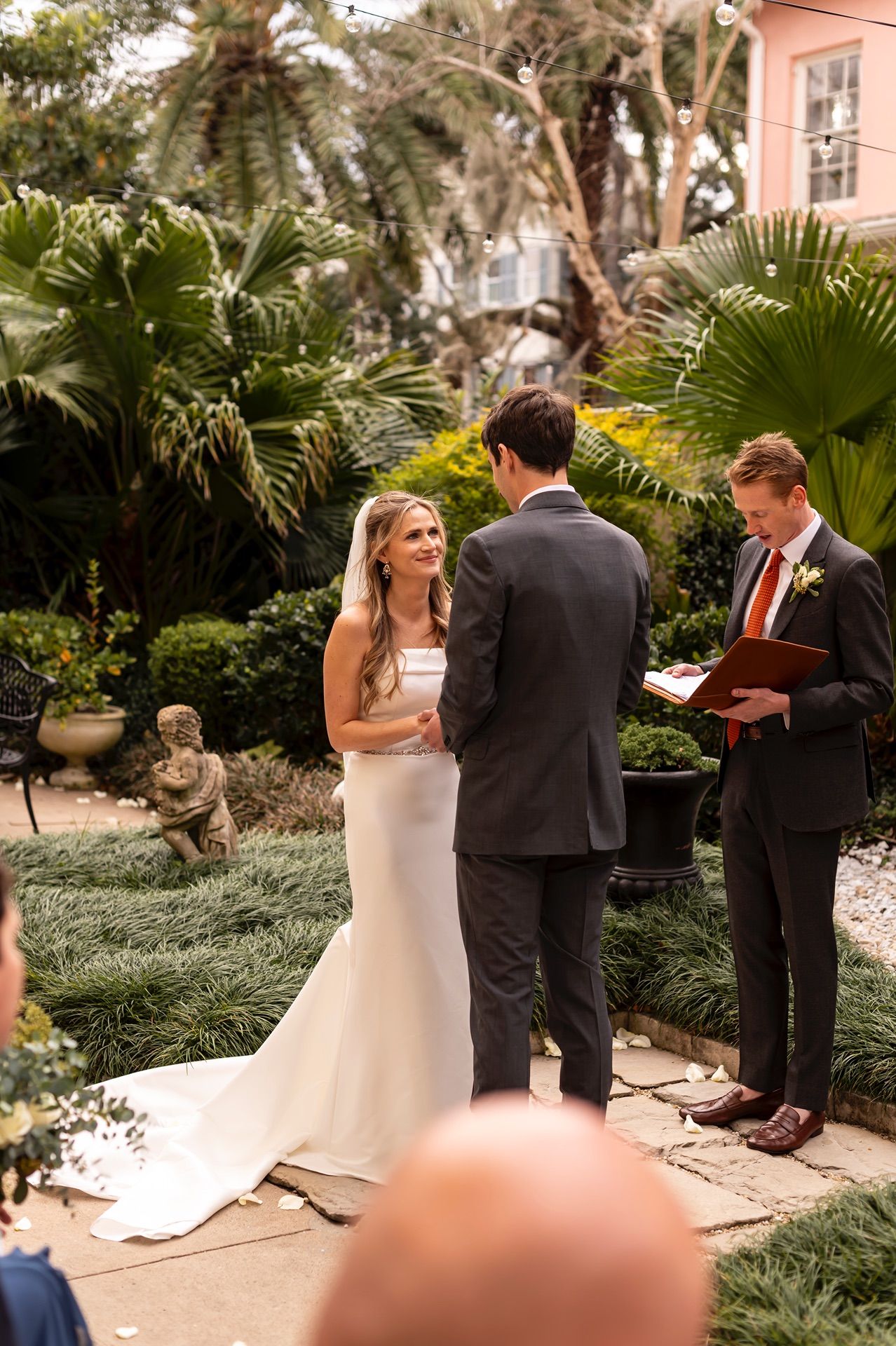 Wedding ceremony taking place in a lush garden setting with tropical palm trees and pink stucco walls.