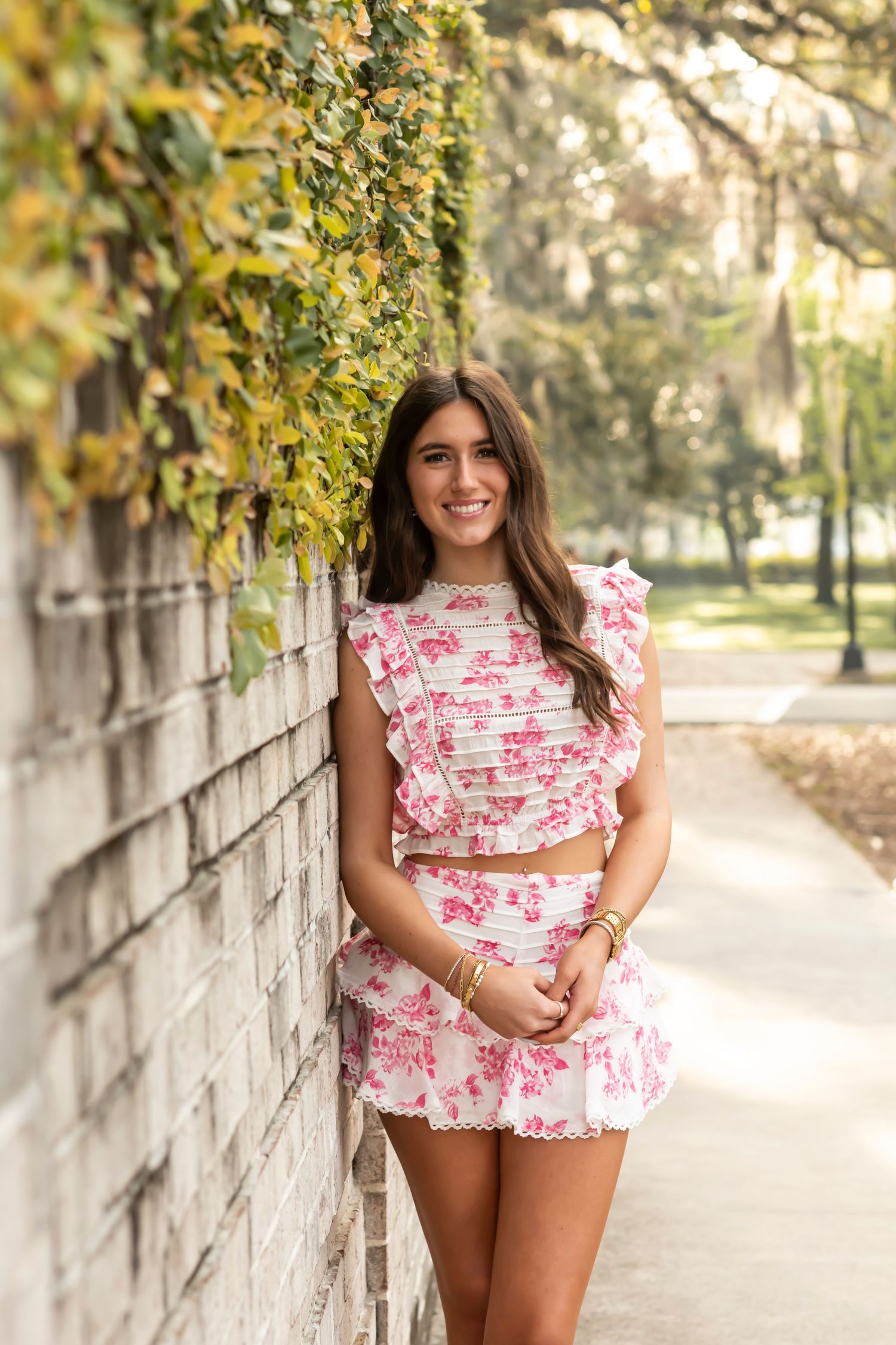 A pink floral ruffle dress creates a romantic silhouette against a sunlit garden pathway with stone walls and greenery.
