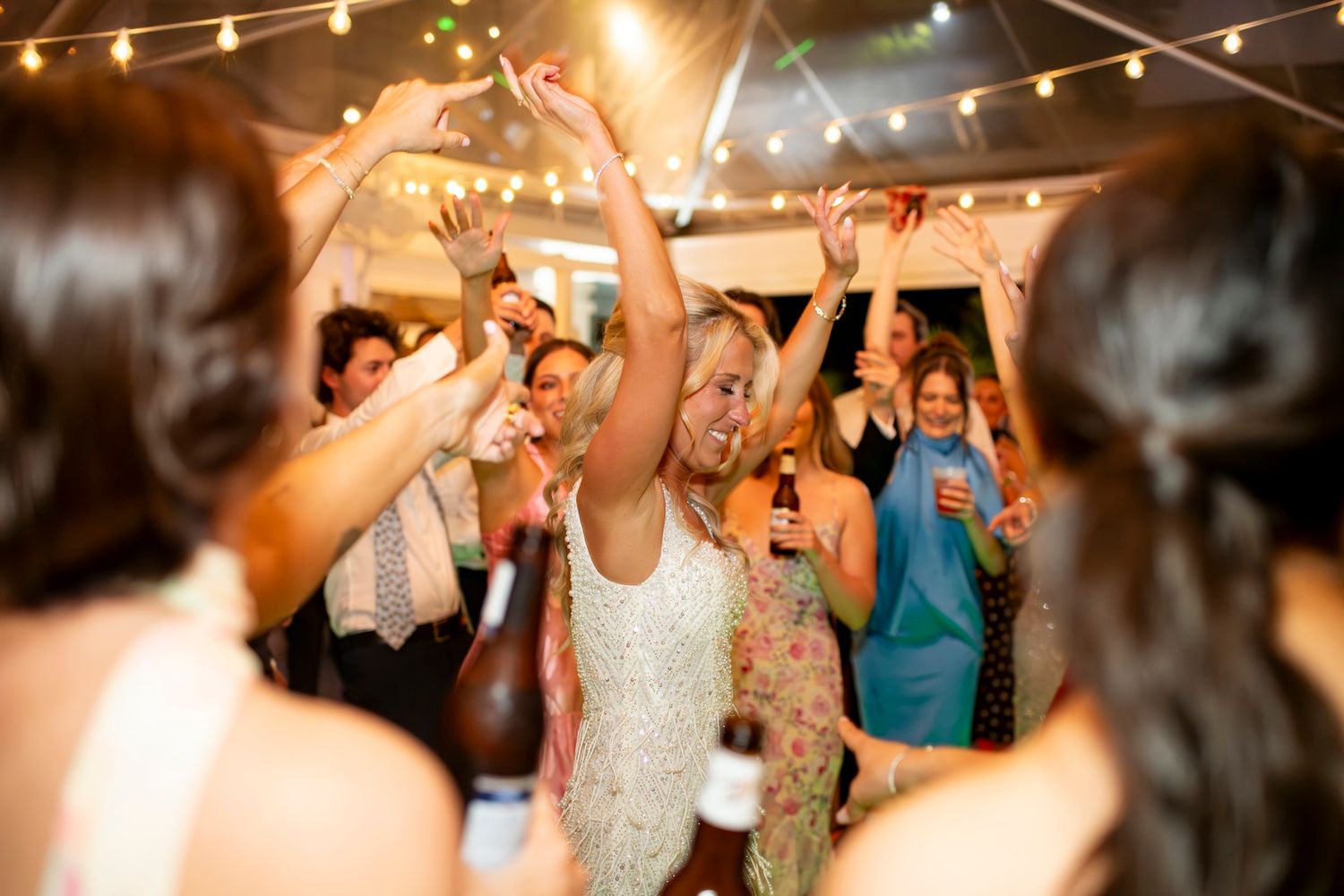 Bride dancing joyfully with guests at a lively wedding reception under warm string lights.