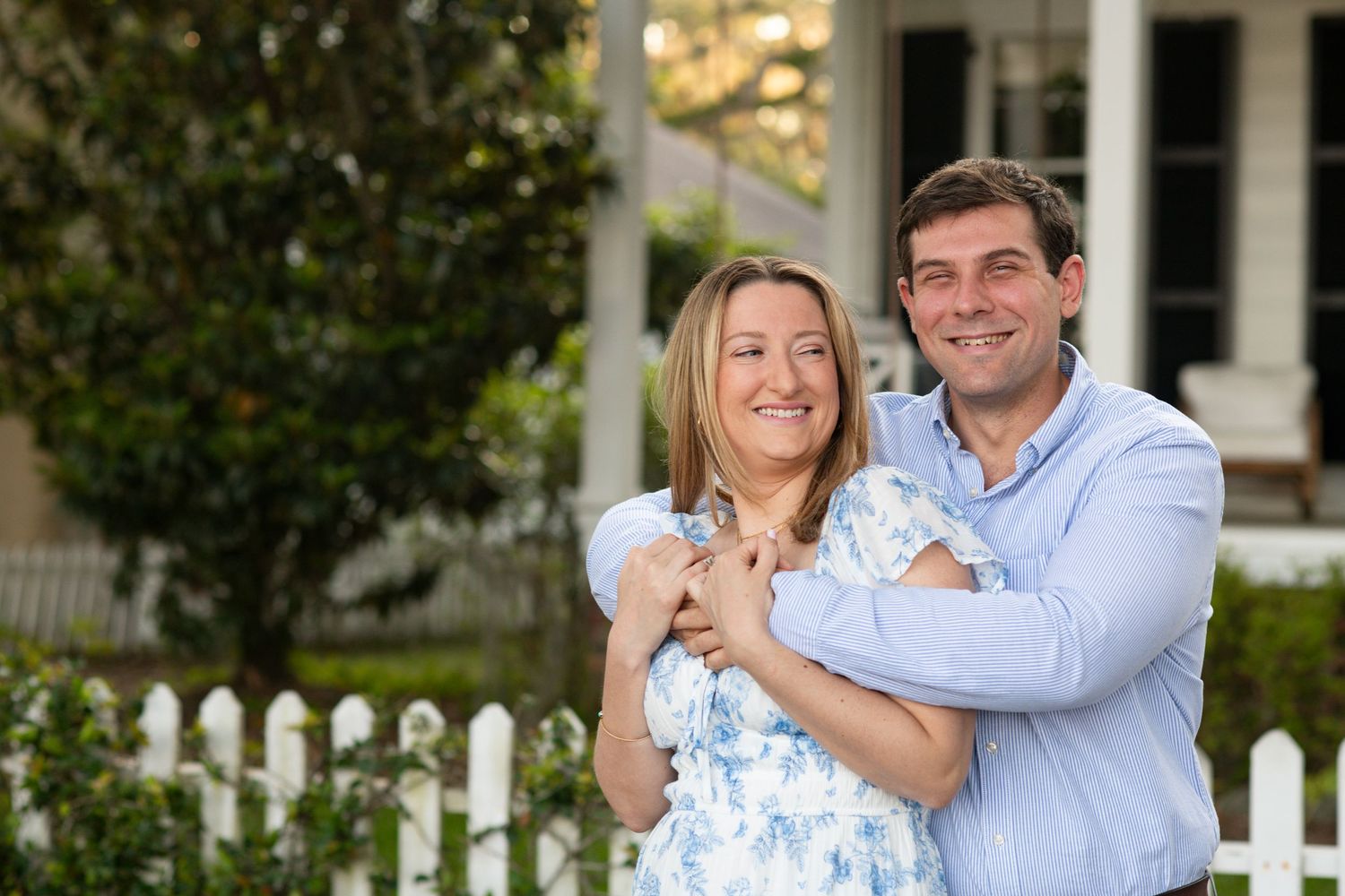 Couple embraces outdoors in front of white picket fence on sunny day wearing coordinated blue and white outfits.