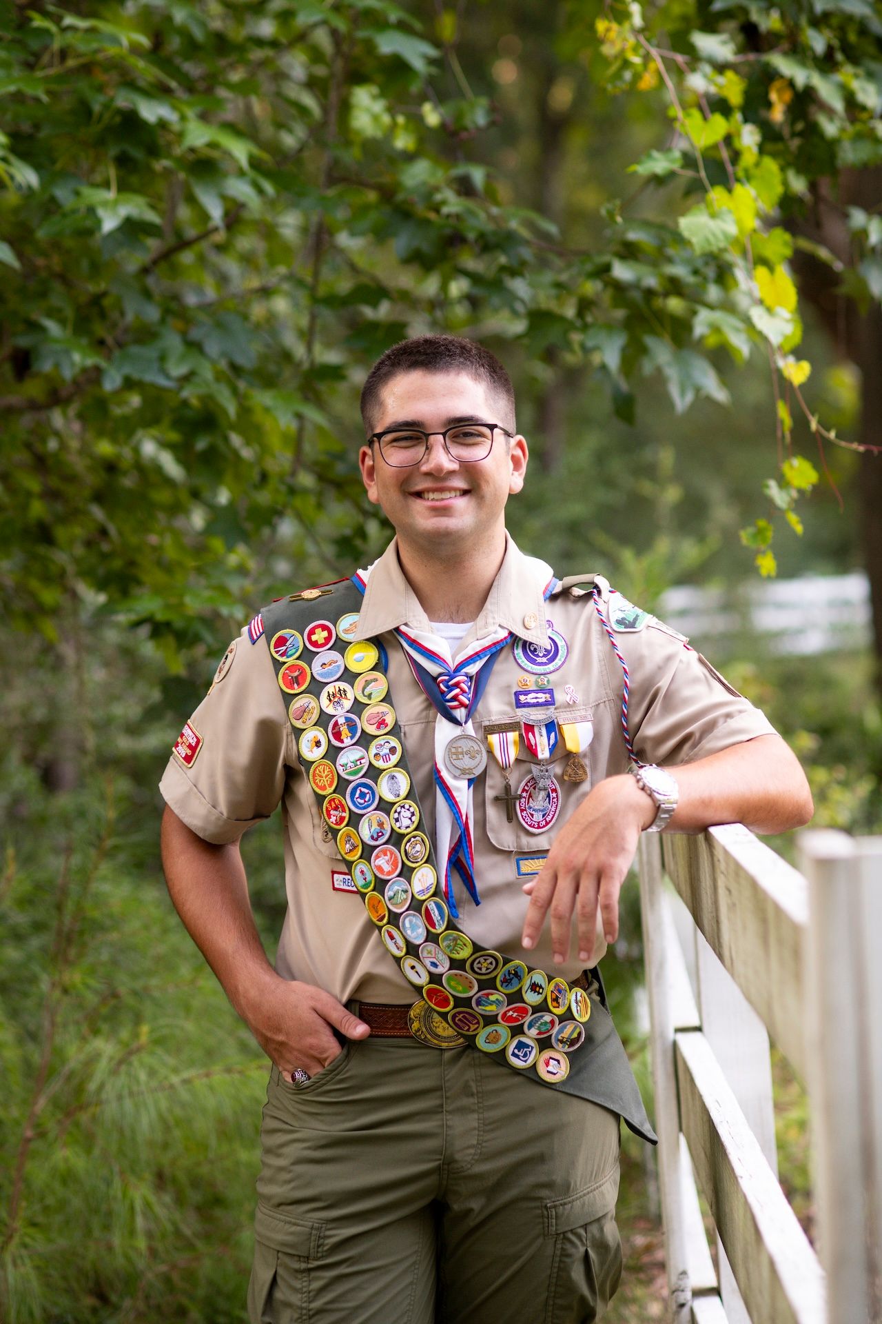 Eagle Scout in uniform with merit badge sash leans against white fence at Holly Oaks on the Marsh for his senior portraits.