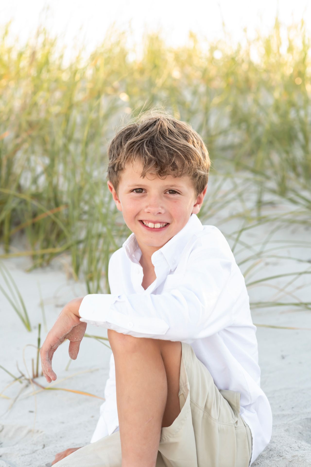 Young child in white button-down shirt sits on sandy beach with sea grass in warm golden sunlight.