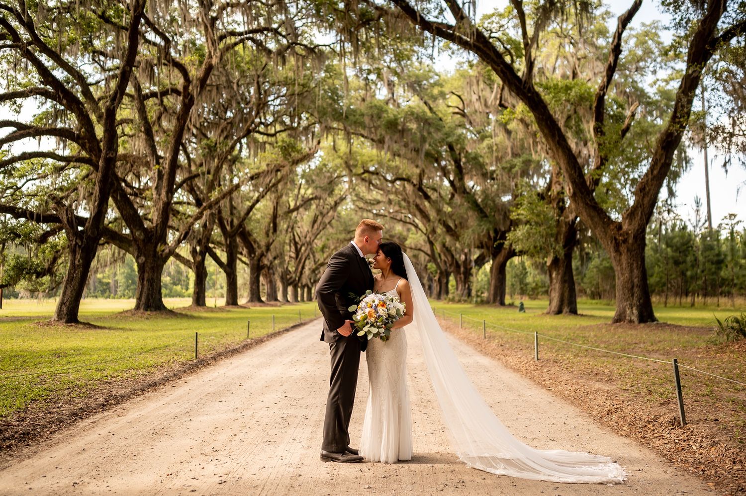 A tree-lined pathway creates a romantic setting for wedding photos in a lush Southern garden estate.