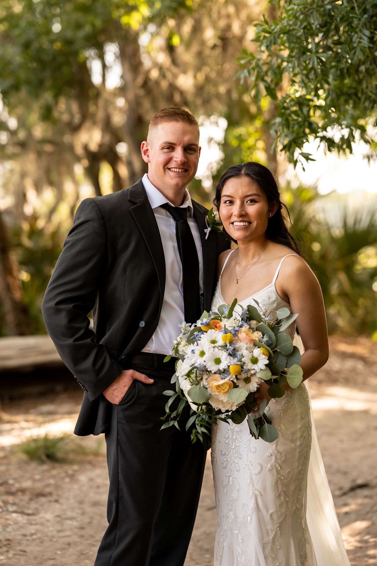 A couple poses outdoors in wedding attire with a beautiful bouquet of white and yellow flowers amongst natural foliage.