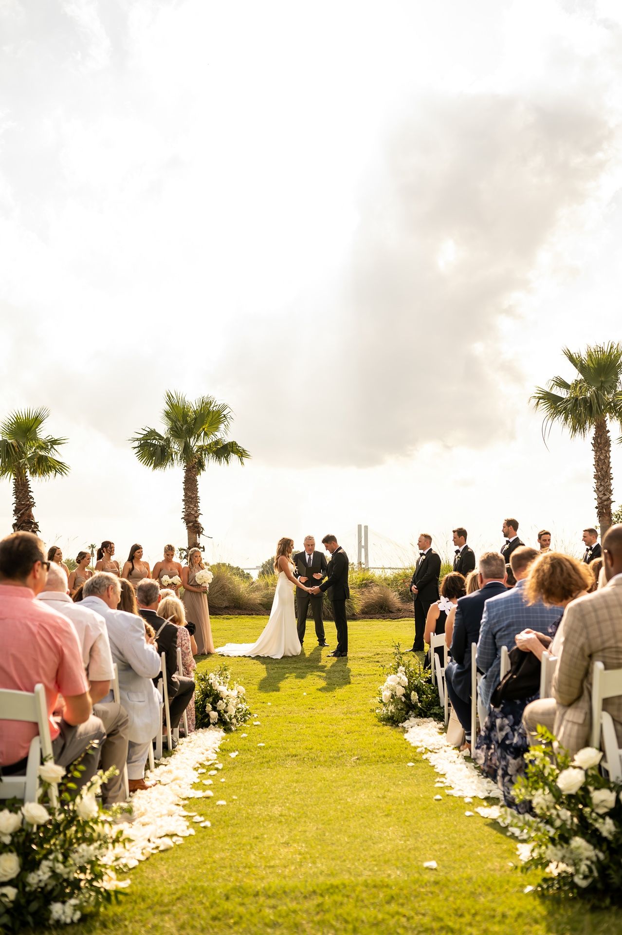 Outdoor beach wedding ceremony surrounded by palm trees and white floral arrangements at sunset.