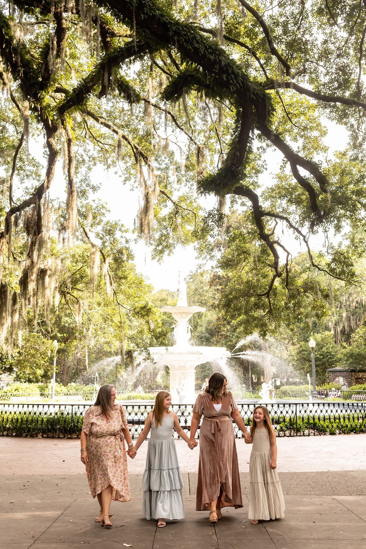 Four people in period costumes walking along a white picket fence under moss-draped oak trees at a historic site.