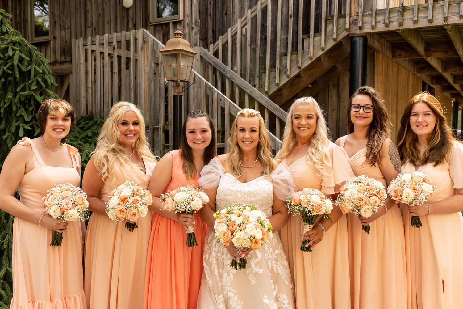 Wedding party wearing peach colored dresses pose together holding bouquets against rustic wooden backdrop.