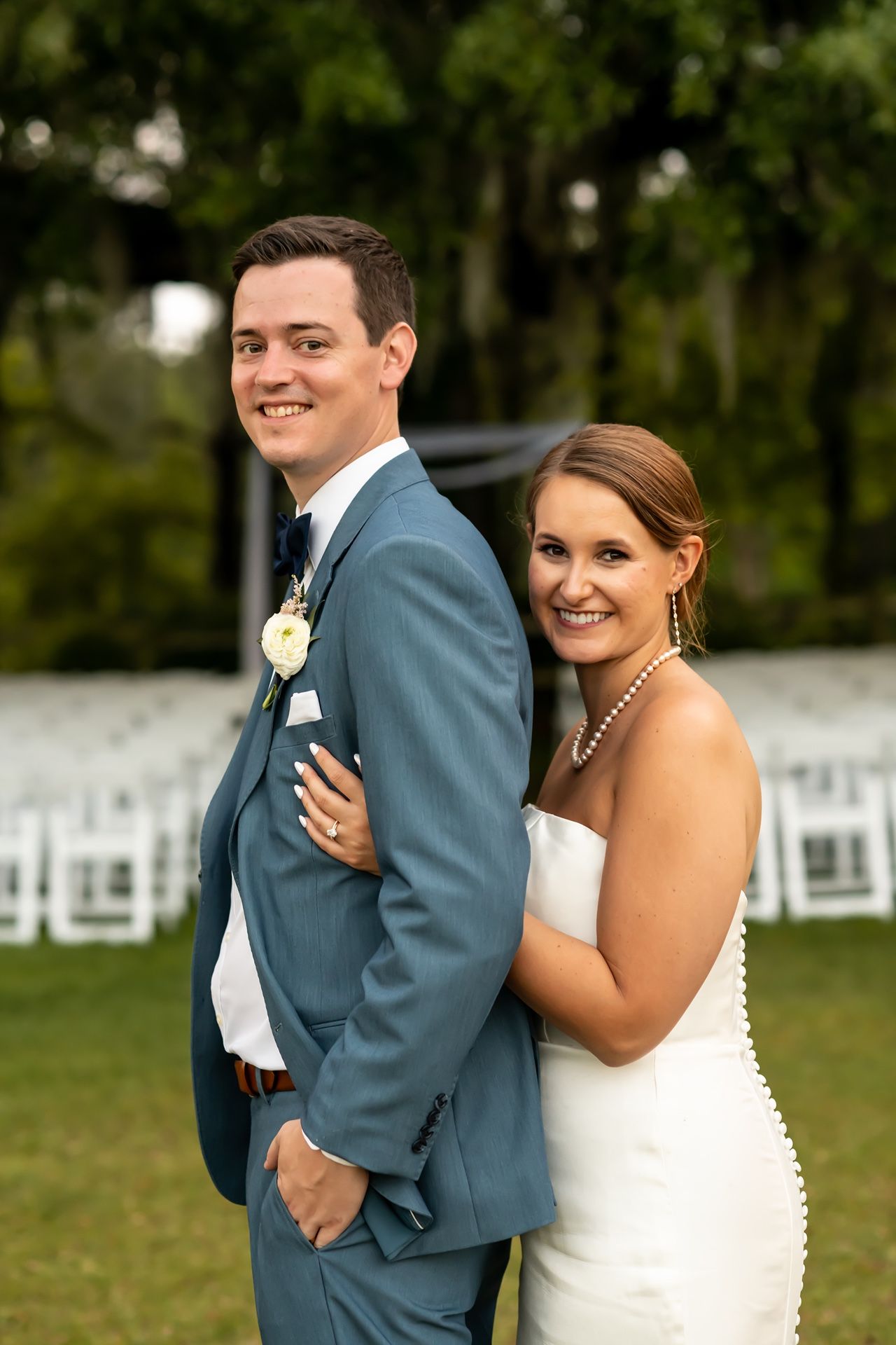 A bride and groom pose together outdoors on their wedding day, with white chairs and greenery in the background.