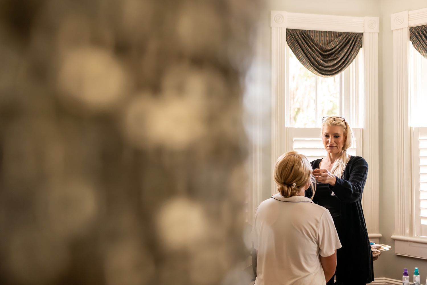 Two people stand in a bright corner room with elegant draped window treatments during wedding preparation.