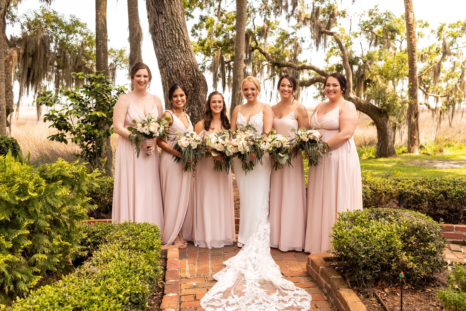 Five bridesmaids in blush pink dresses stand with bouquets on a brick path surrounded by Spanish moss and garden landscaping.