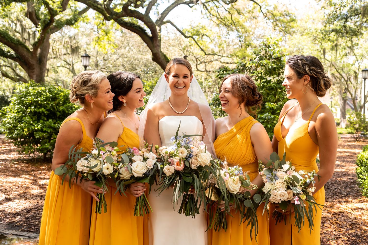 Wedding party wearing bright yellow dresses and holding lush greenery bouquets pose together outdoors in sunlit garden setting.