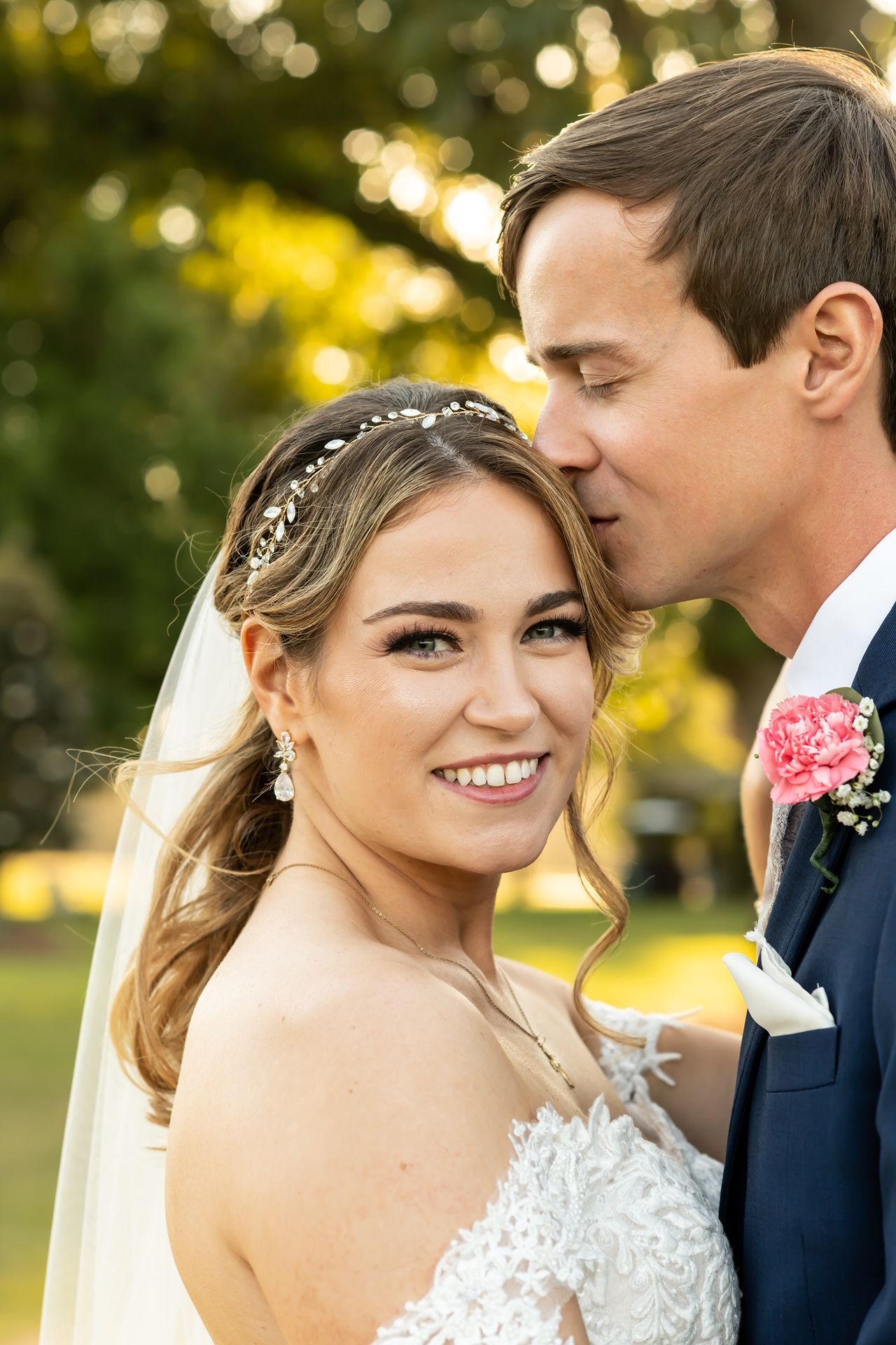 Bride wearing lace wedding dress and veil smiles while groom in navy suit leans in during outdoor portrait session at sunset.