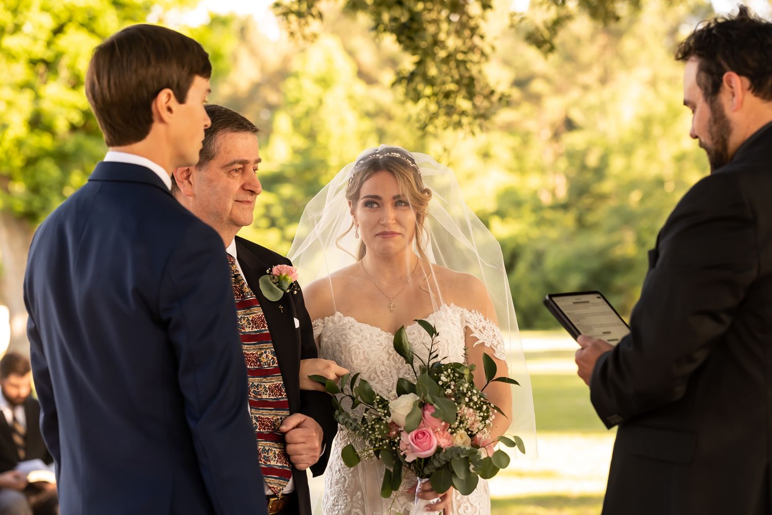 A couple exchanges vows during an outdoor wedding ceremony with lush greenery and golden sunlight in the background.