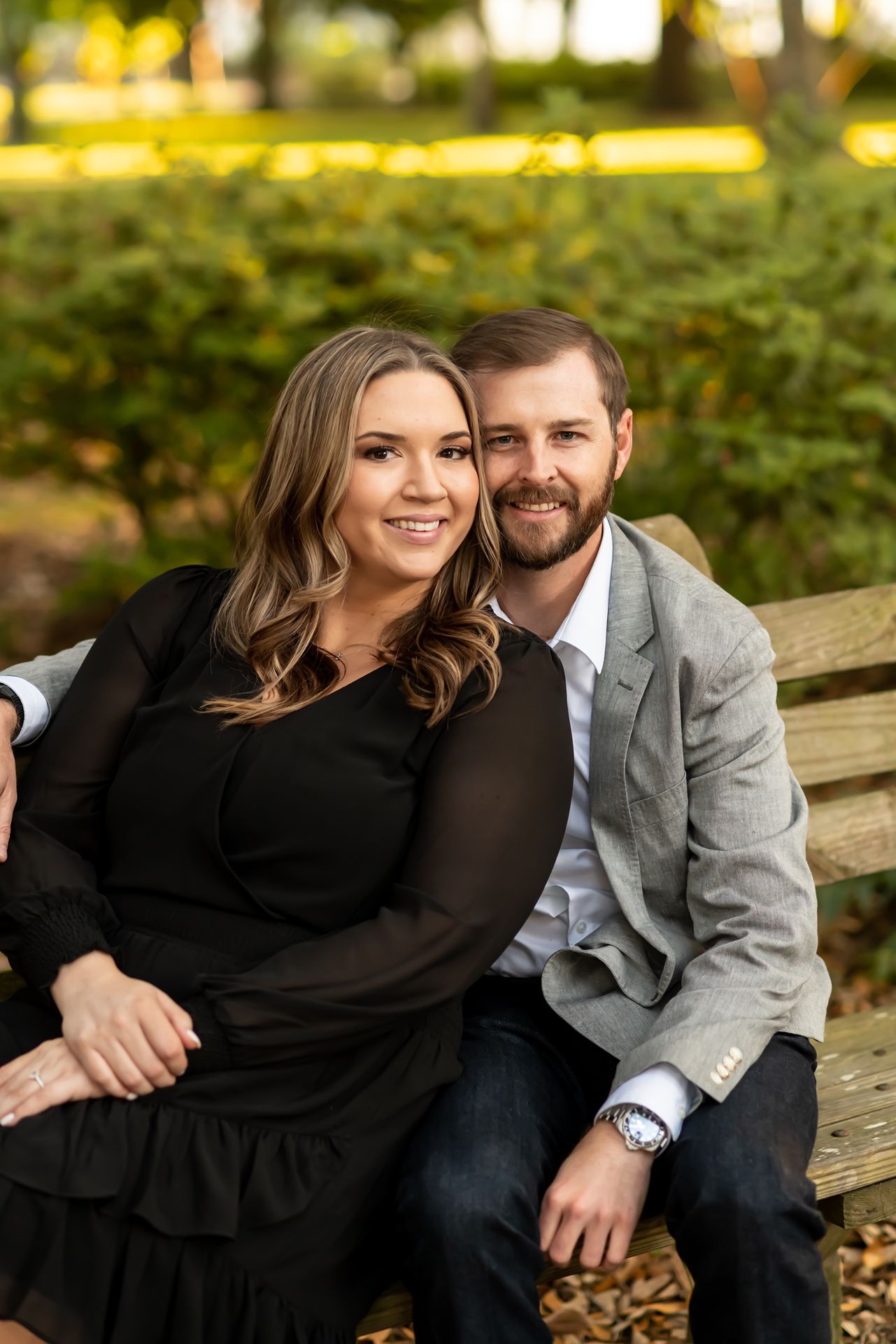 A couple sitting together on a wooden bench outdoors with green foliage and warm lighting in the background.