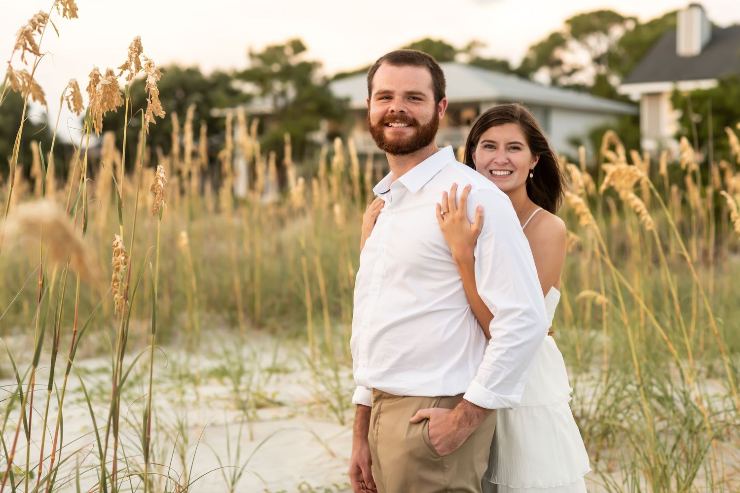 A couple shares joyful moments together on a sandy beach with tall seagrass at golden hour.
