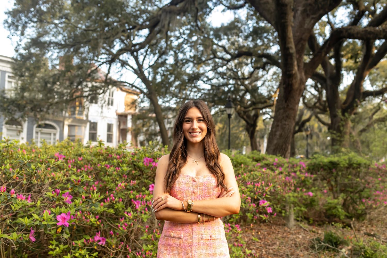A smiling woman in coral dress poses outdoors among pretty purple flower gardens and oak trees.