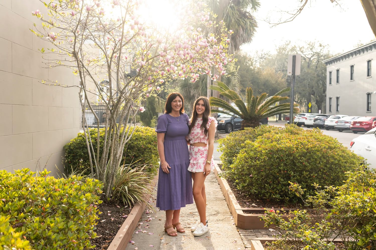 Two people standing together on a garden pathway with yellow flowers and palm trees in bright sunlight.