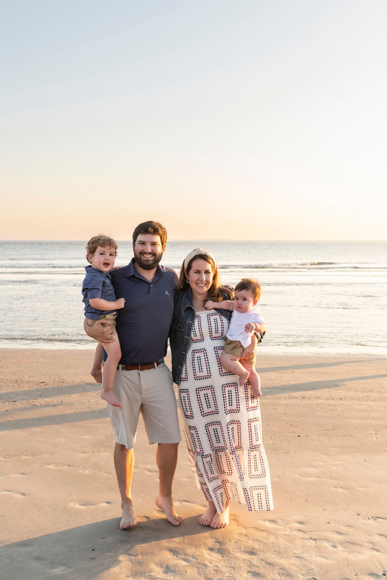 Family of four poses on a sandy beach during golden hour with ocean waves in the background.