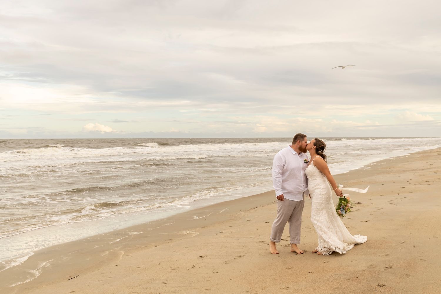 A couple in wedding attire shares an intimate moment while walking along a misty ocean beach at sunset.