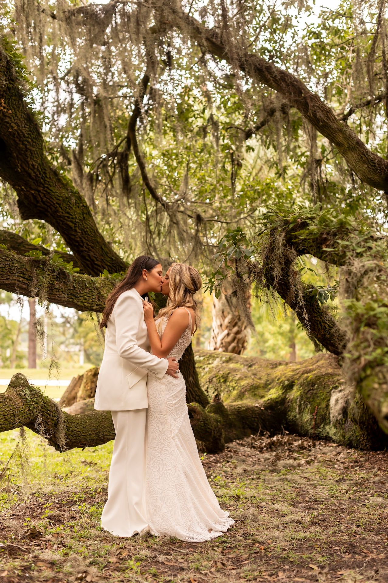 A couple in white wedding attire share a romantic moment under a majestic Spanish moss-draped oak tree.