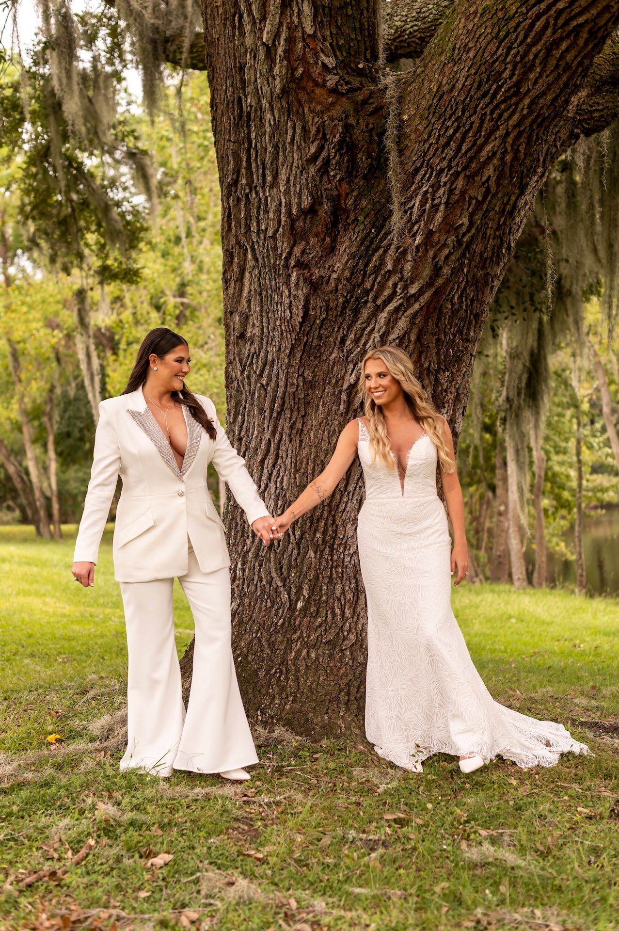 A romantic wedding photo of two brides in white outfits holding hands by a large oak tree in a sunlit park.