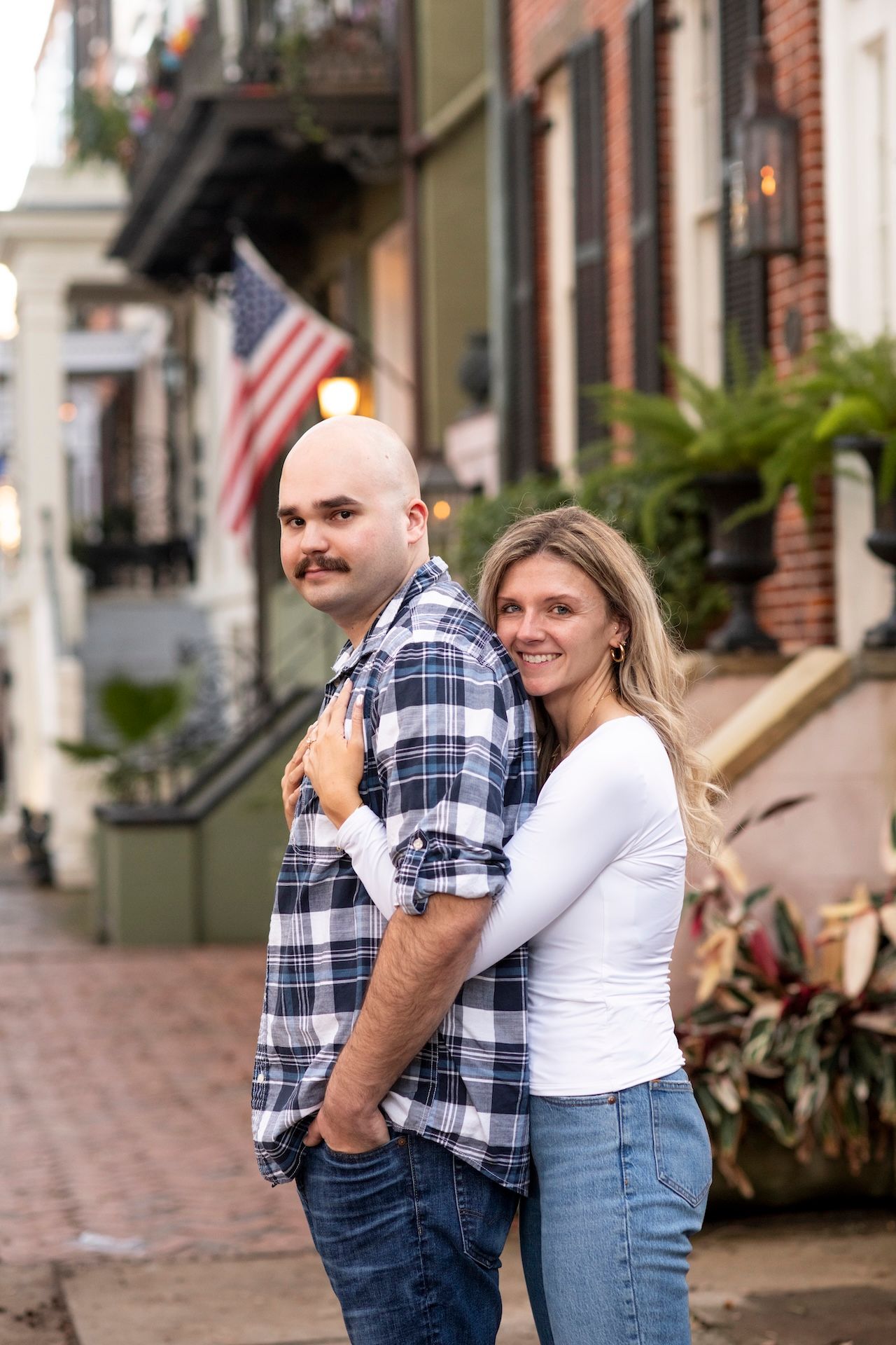 A couple embraces on a charming brick sidewalk in front of historic townhouses with an American flag in the background.