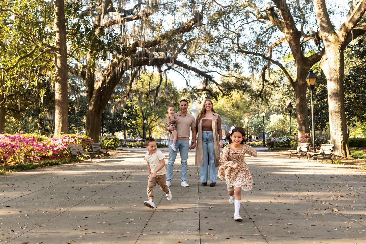 A happy family walks along a tree-lined path with pink azalea bushes in a sunny spring garden.