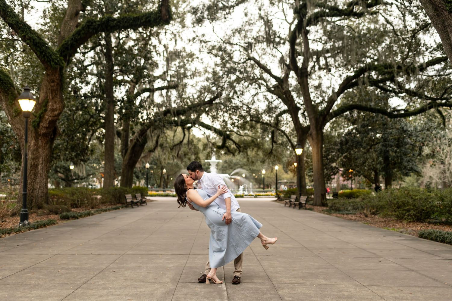 A romantic couple dips in an elegant pose along a tree-lined pathway in a scenic park setting.
