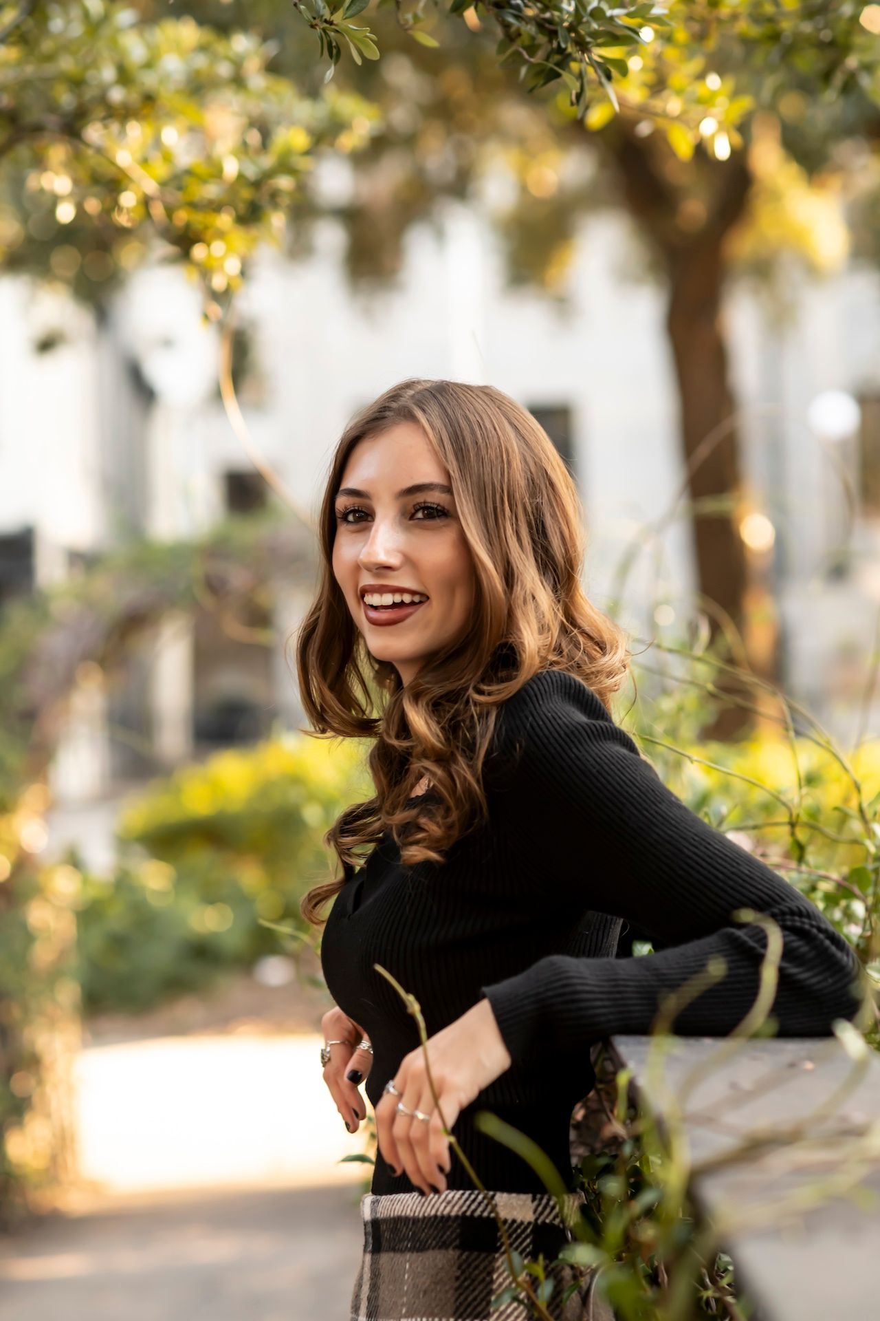 Person in black sweater stands amid autumn foliage with warm sunlit background.