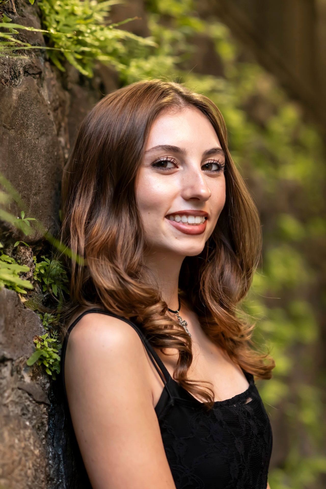 Portrait of someone in a black dress leaning against a tree trunk with lush green foliage in the background.