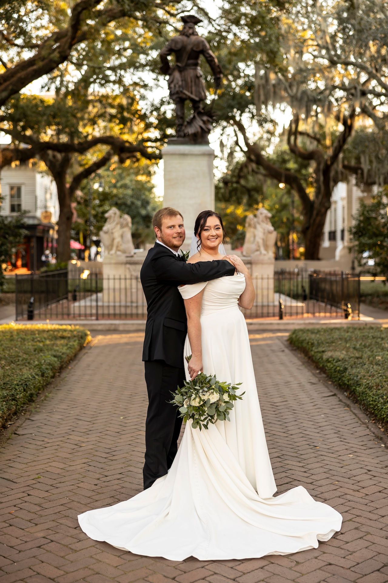 A wedding couple poses together in a romantic outdoor setting with Spanish moss draped oak trees and a historic statue in Savannah.