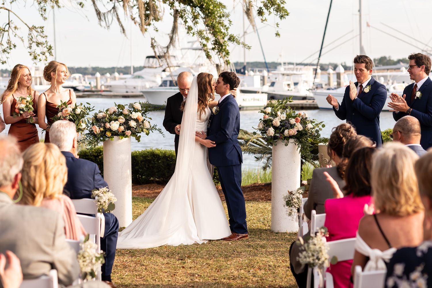 Romantic waterfront wedding ceremony with floral columns and sailboats docked in marina during golden hour.