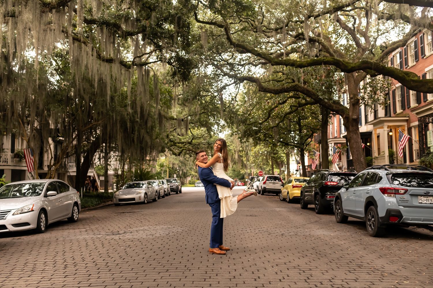 Person stands on cobblestone street lined with Spanish moss-draped trees and parked cars in historic Savannah, Georgia.