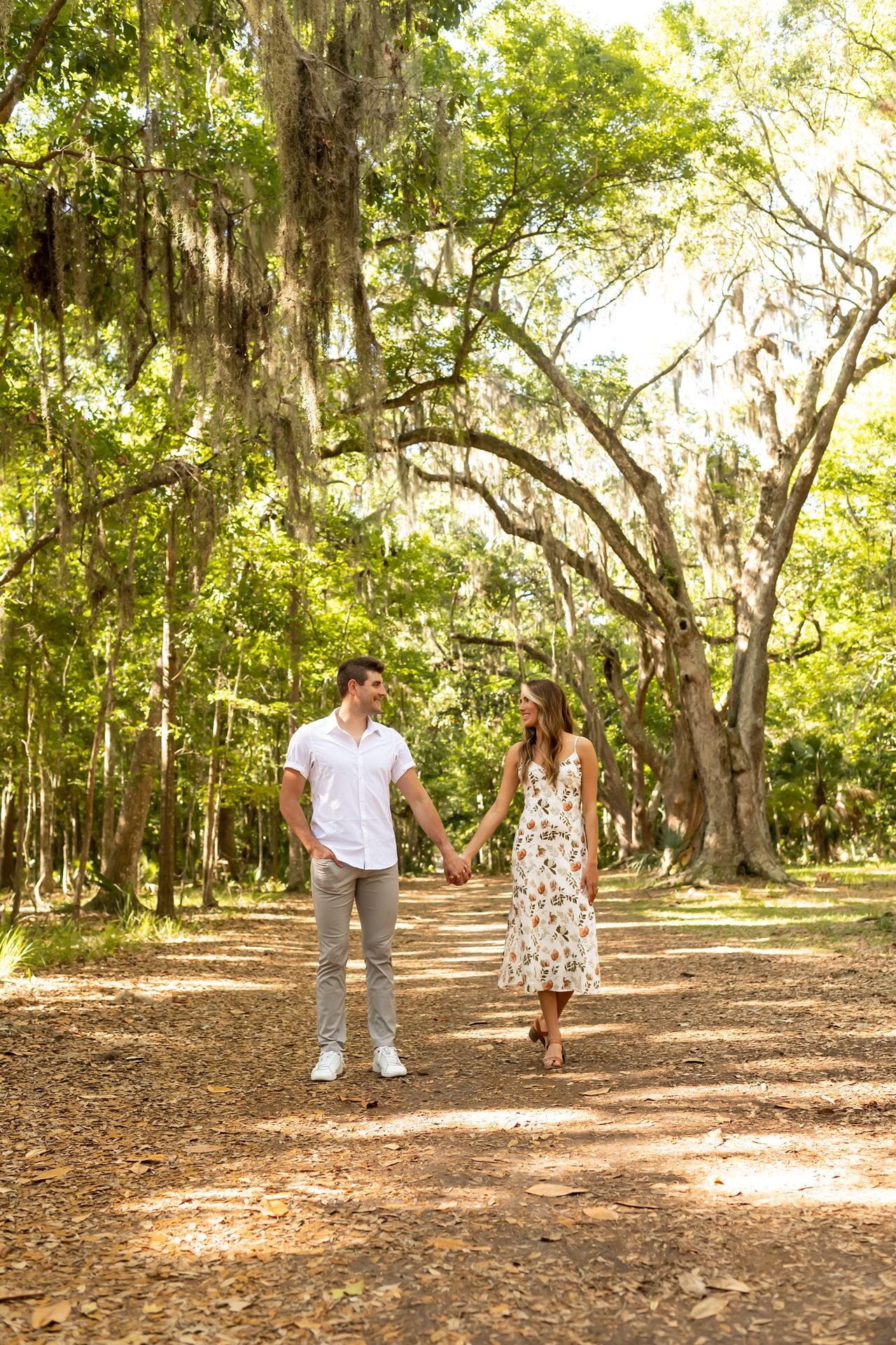 Couple walking hand in hand on a nature trail under Spanish moss-draped oak trees in a sunlit Southern park.