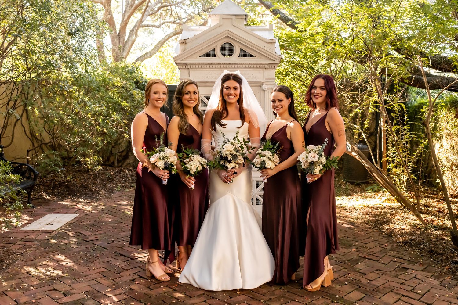 A bride in white gown stands with bridesmaids wearing burgundy dresses in front of a garden gazebo.