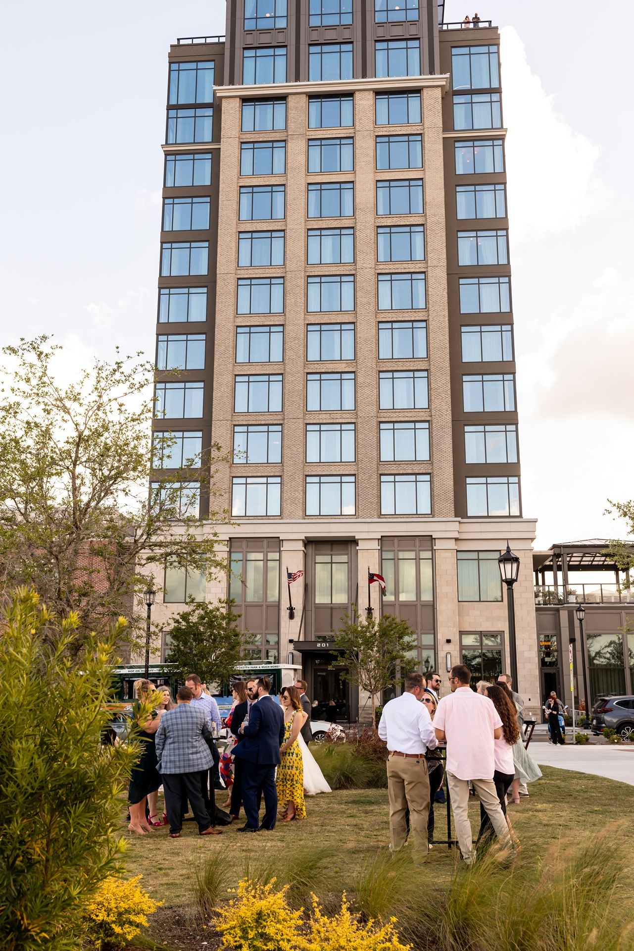 Hotel exterior with guests gathering outside modern high-rise building surrounded by landscaped gardens.