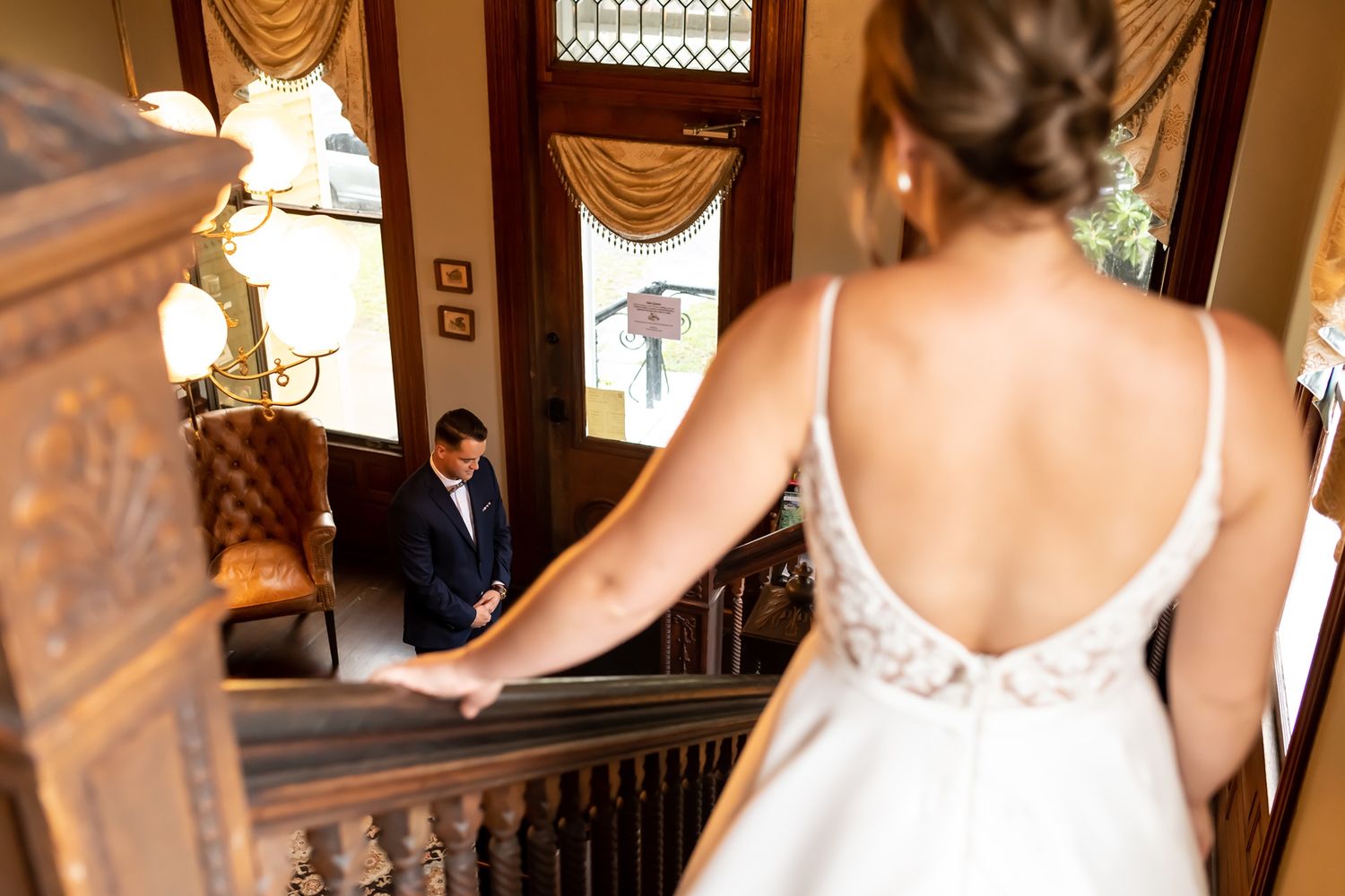 Bride in elegant white lace dress walking up ornate wooden staircase in grand venue with golden drapery.